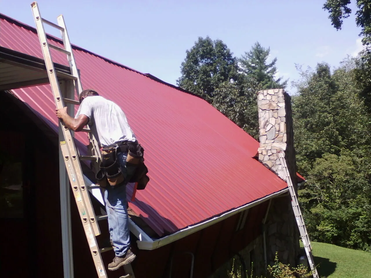 Roofer on a ladder beside a red metal roof and stone chimney — Soffit & Fascia Repair by Roofing Brothers in Travis Afb